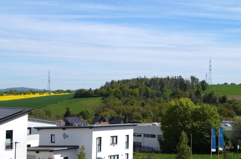 White modern building in the foreground overlooking green fields and a distant tree-covered hill under a blue sky with power pylons across the horizon.