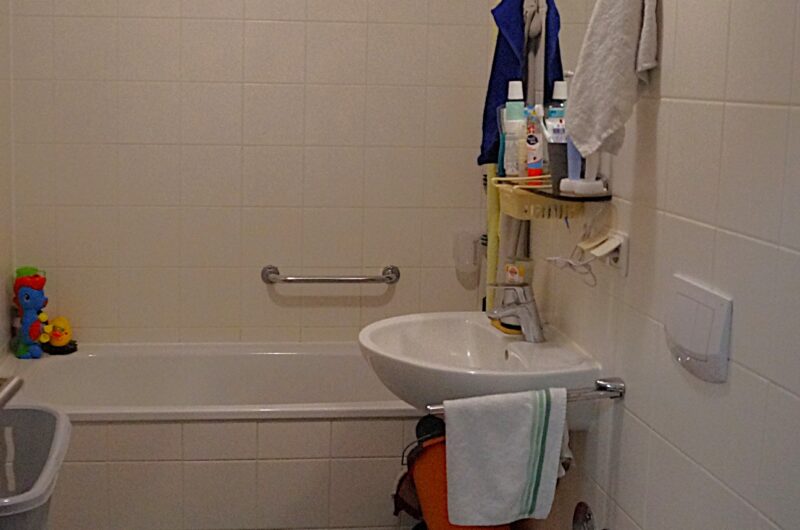Small bathroom with a bathtub, sink, and toilet; blue mats on a brown tiled floor and towels hanging above a shelf.