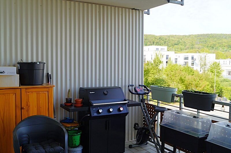 Balcony or covered porch with a black gas grill, wooden cabinet, plant pots, and outdoor furniture against a corrugated metal wall; view to trees and modern buildings beyond.