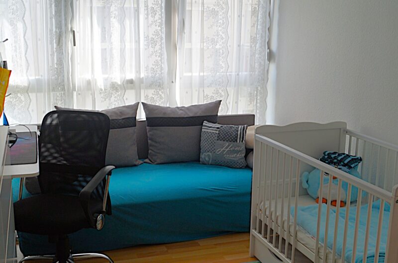 Bright children's room with a white crib, blue couch, and lace curtains by large windows; study desk and black office chair nearby.