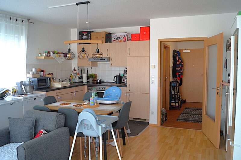 Open-plan kitchen and dining area with light wood cabinets, pendant lights, and a wooden dining table surrounded by chairs; doorway to a hallway with coats hanging.