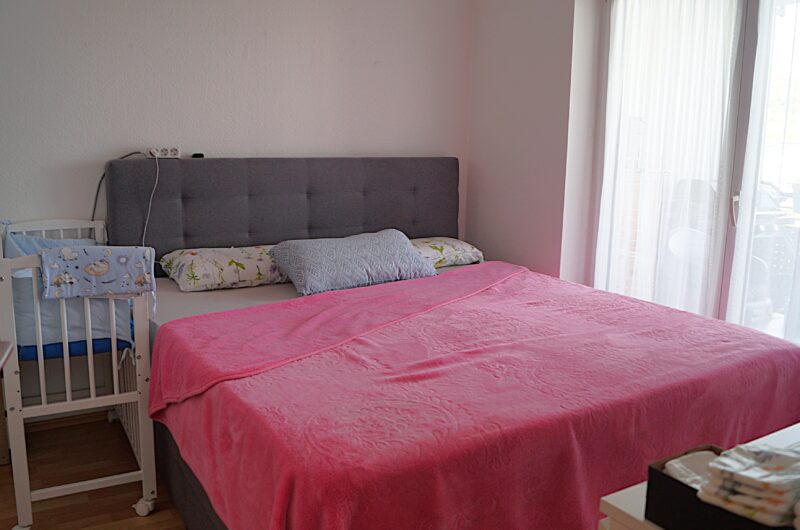 Bedroom with a pink quilted bed, gray upholstered headboard, and a white crib on the left next to a wall outlet and cords.