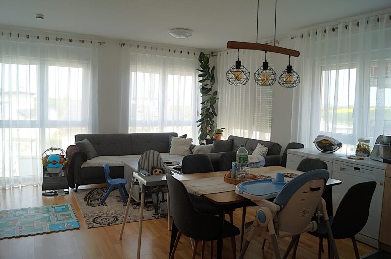 Bright living room with white sheer curtains, a gray sofa, and three geometric pendant lights over a wooden table outside a windowed wall