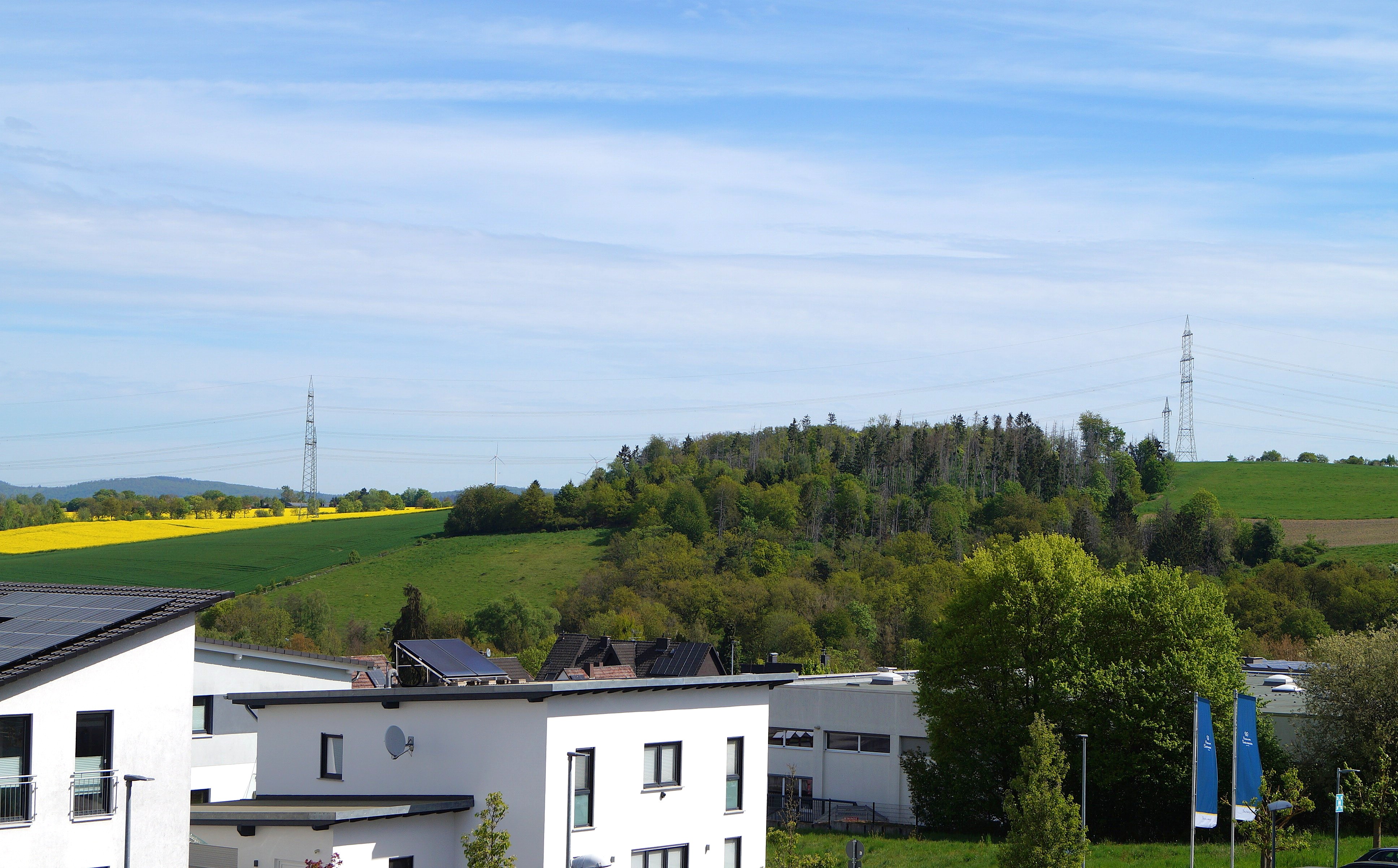 White residential buildings in the foreground with green fields, trees and distant electric pylons under a blue sky. - Informative scene of a rural-urban edge and power lines.