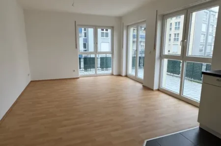 Empty modern living room with light wood flooring, white walls, and large windows leading to a balcony.