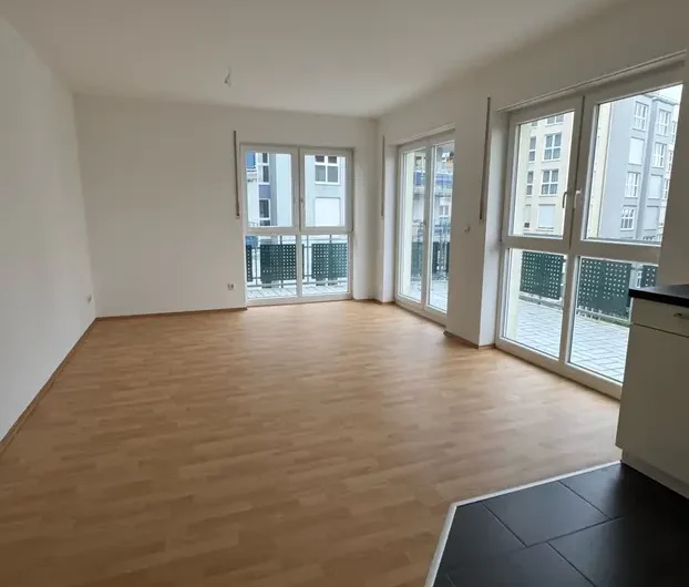 Empty modern living room with light wood flooring, white walls, and large windows leading to a balcony.