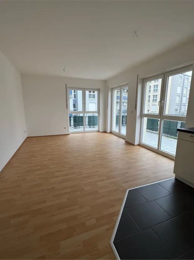 Empty modern living room with light wood flooring, white walls, and large windows leading to a balcony.