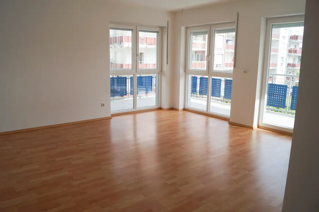 Bright empty living room with warm wood flooring and large windows opening to a balcony view outside.