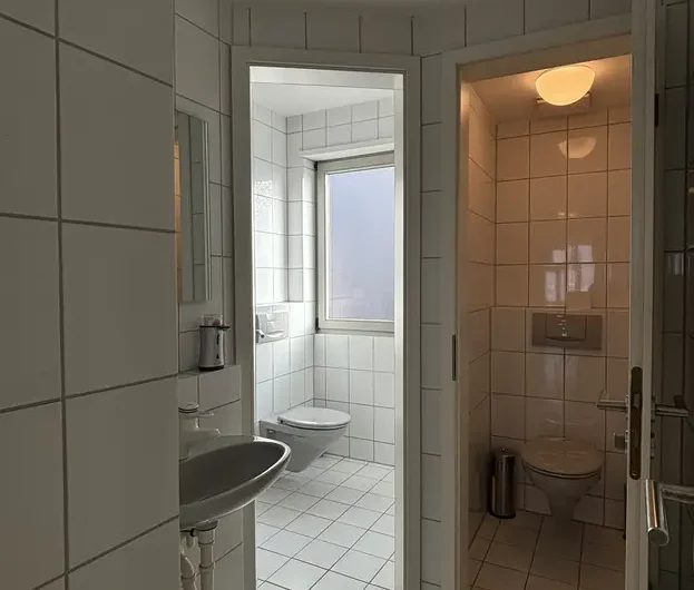 White tiled bathroom with a wall-mounted sink in the foreground, a toilet by a window straight ahead, and a separate toilet cubicle to the right.