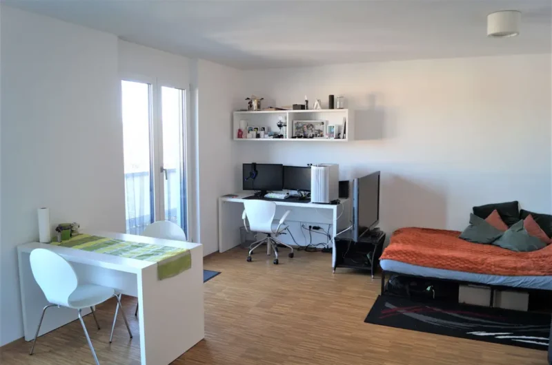 Bright studio with a white desk and dual monitors, computer tower, and a bed with an orange blanket on the right.