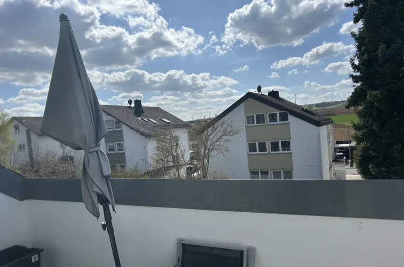 Balcony with a folded grey patio umbrella; white wall, and a view of neighboring apartment buildings under a blue, cloudy sky.