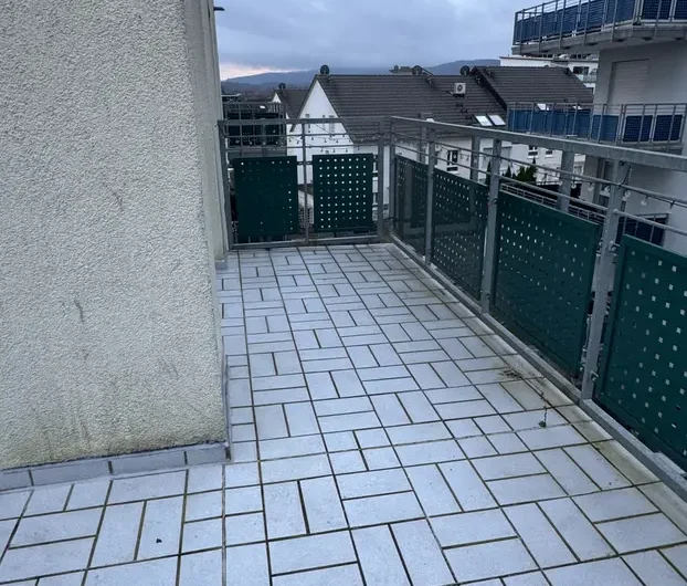 Balcony with light gray tile floor and green perforated railing, overlooking neighboring rooftops under a cloudy sky.