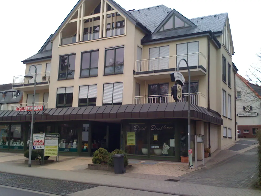 Three-story beige building with large windows and ground-floor shops under a brown awning on a cobblestone street.