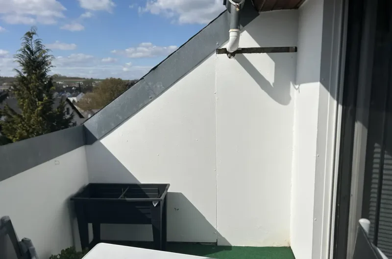 Small rooftop balcony with white walls, a black grill against the left wall, a white table in the foreground, and green outdoor flooring; blue sky above.