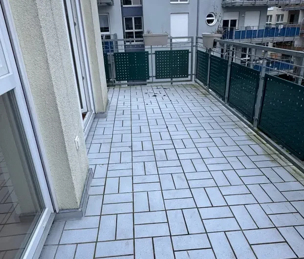 Apartment balcony with light gray patterned tiles, glass and metal railing, overlooking other building balconies in a city block.