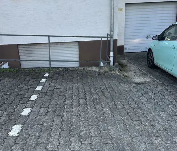 Light blue car parked beside a white building with a metal railing, on a cobblestone driveway near a garage door.
