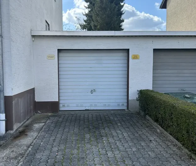 Narrow cobblestone driveway leading to a white garage door, with a hedge on the right and a tall evergreen tree against a blue sky behind the building.