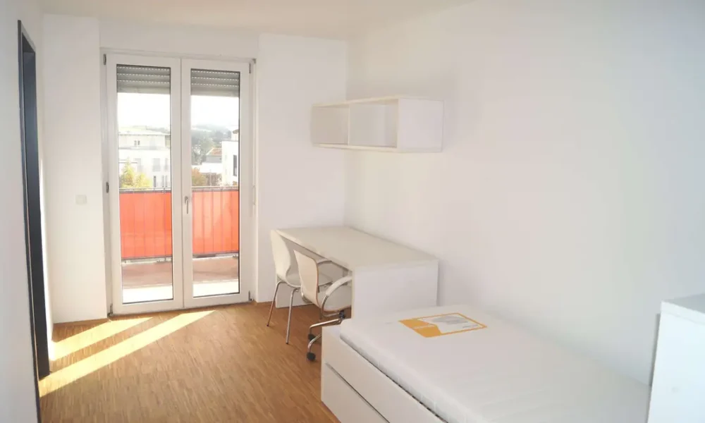Bright white bedroom with a single bed, desk, and wall shelf; glass balcony doors open to a reddish railing outside.