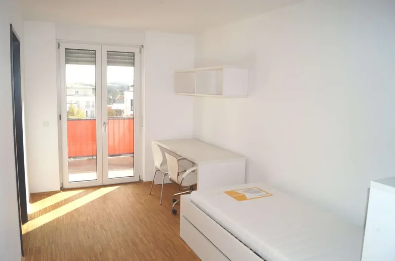 14 - DEBUS Immobilien Bright white bedroom with a single bed, desk, and wall shelf; glass balcony doors open to a reddish railing outside.