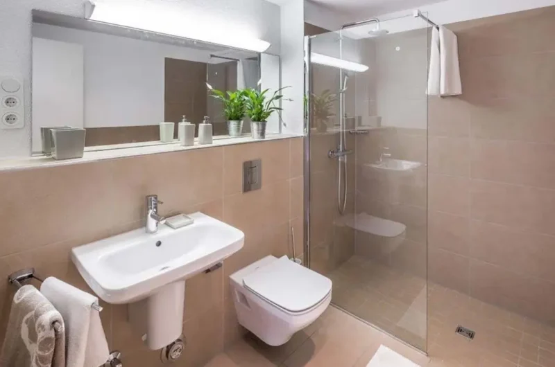 Modern beige-tiled bathroom with a wall-mounted toilet, pedestal sink, and glass-enclosed shower.
