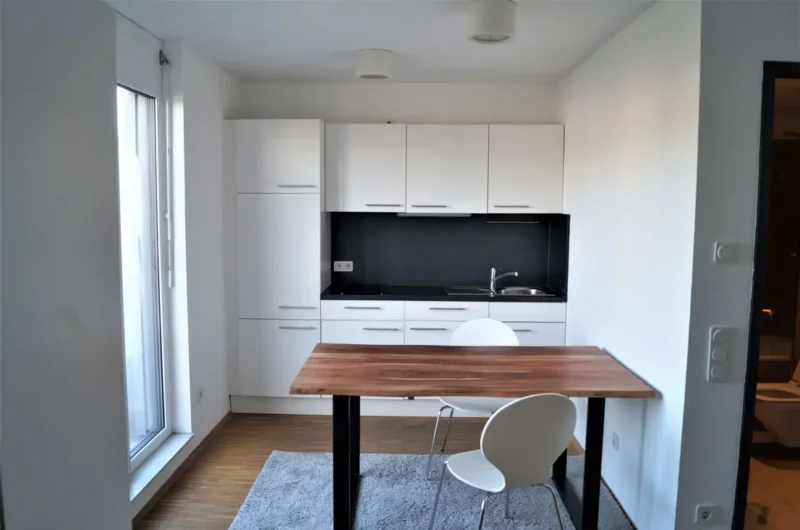 Modern white kitchen with a black backsplash, wooden dining table, and white chairs near a large window.