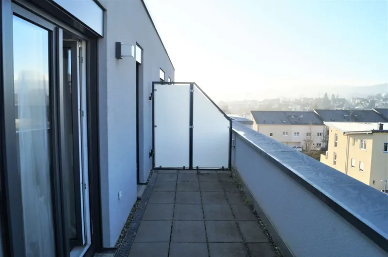 Long balcony on a gray apartment building with a frosted glass divider at the far end, overlooking a cityscape.