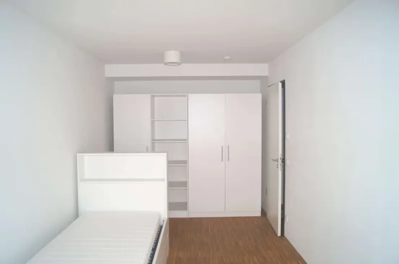 White minimalist bedroom with a single bed, open-shelving unit, and a large white wardrobe against light walls and a wooden floor.
