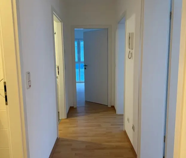 Narrow white hallway with a light wood floor and several closed doors, ending with an open door to a bright room.