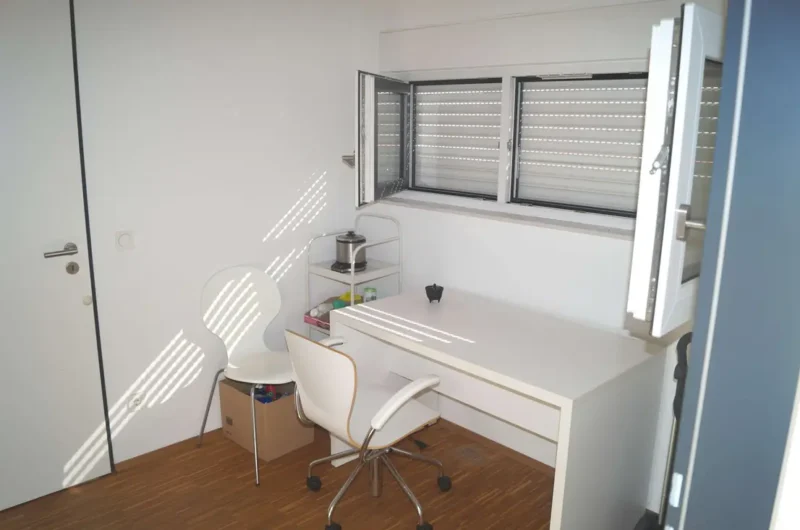 Small white home office with a desk, rolling chair, and a shelving cart; sunlight streams through horizontal blinds casting stripes on the wall.