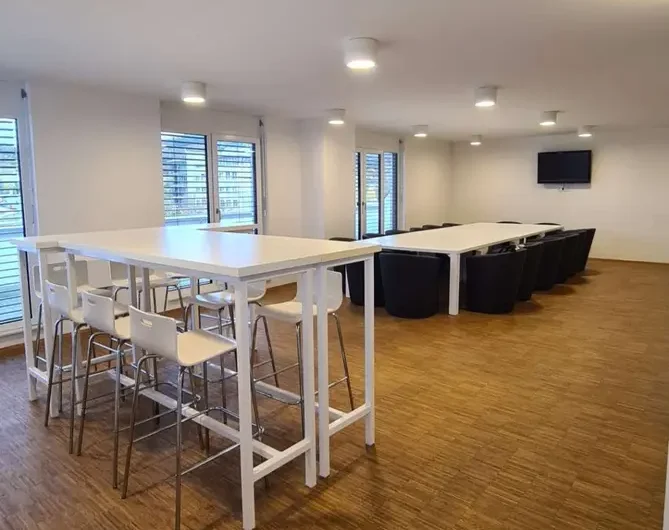 Modern meeting room with long white tables, white stools at a high table, and black armchairs along a second table row; wooden floor and windows along the walls.