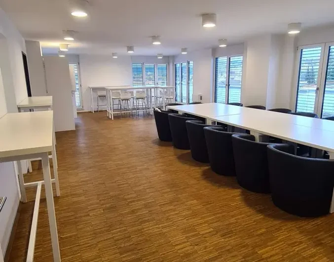 Bright, modern conference room with long white tables, black chairs, and wood-like flooring, large windows with blinds in the background.