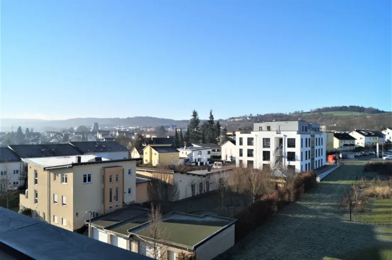 Wide view of a suburban town with modern white and beige apartment buildings under a clear blue sky, with a grassy park area in the foreground.