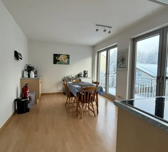 Open-plan dining area with a wooden floor, a rectangular table and six chairs, a coffee station against the left wall, and large glass doors to a balcony on the right.