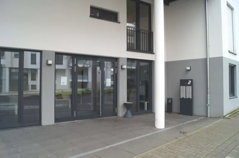 Glass double doors at a modern building entrance, with a white column and a two-tone gray facade; mailbox cabinet marked '2' on the right.