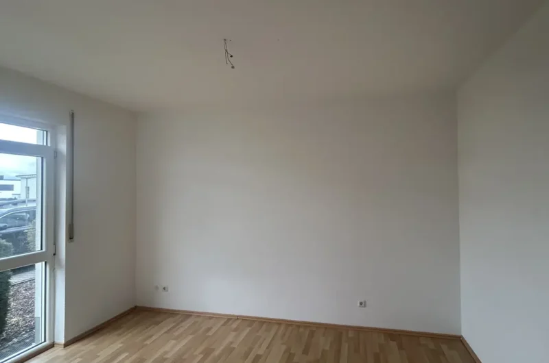Empty white-walled room with light wood floor, a large glass door/window on the left, and hanging electrical wires from the ceiling.