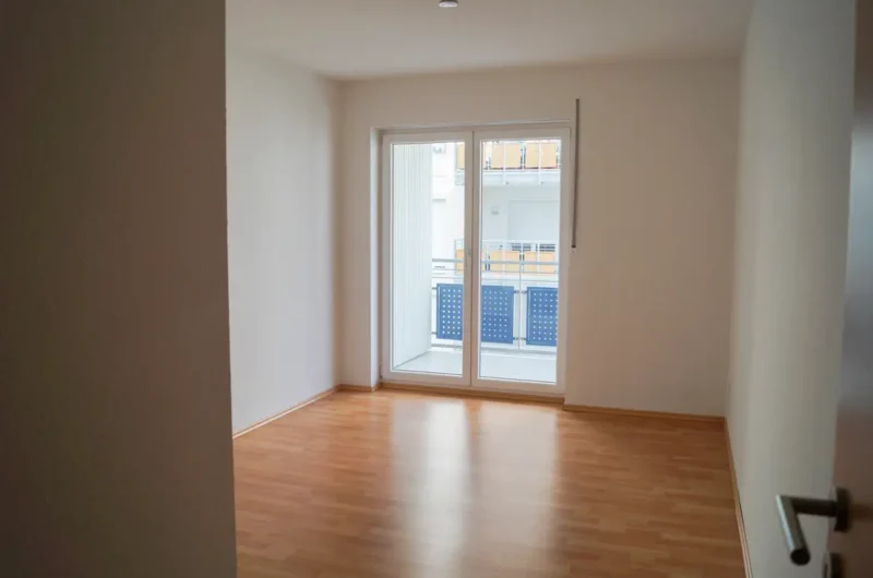 Empty rectangular room with light wood flooring and white walls, sliding glass door to a balcony.