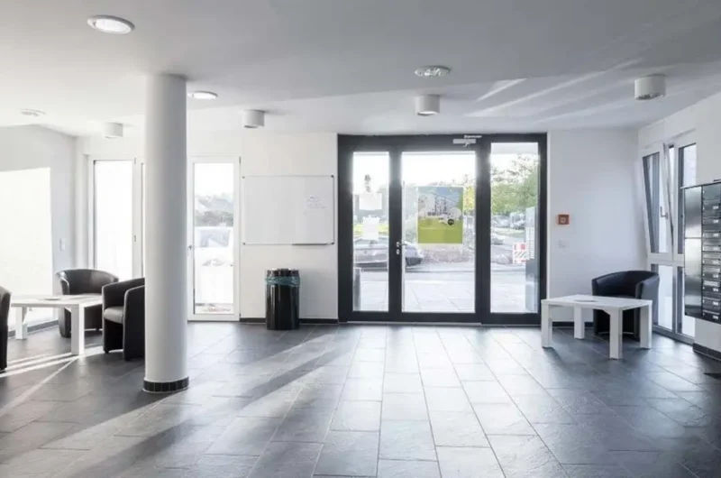 Modern lobby with gray tiled floor, white walls, and a seating area by glass doors.