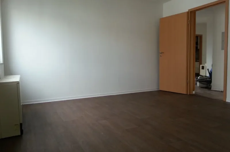 Empty room with dark wood laminate flooring and white walls; a partially open wooden door reveals a adjacent area with cleaning equipment visible.