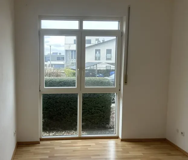 Empty room with a light wood floor and a large white-framed window letting in daylight; hedges visible outside.