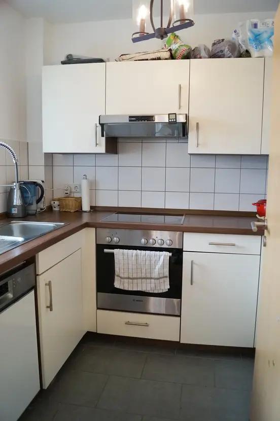 Compact L-shaped kitchen with white cabinets, stainless oven and range, brown countertops, tiled backsplash, sink on the left, and a towel hanging from the oven handle.