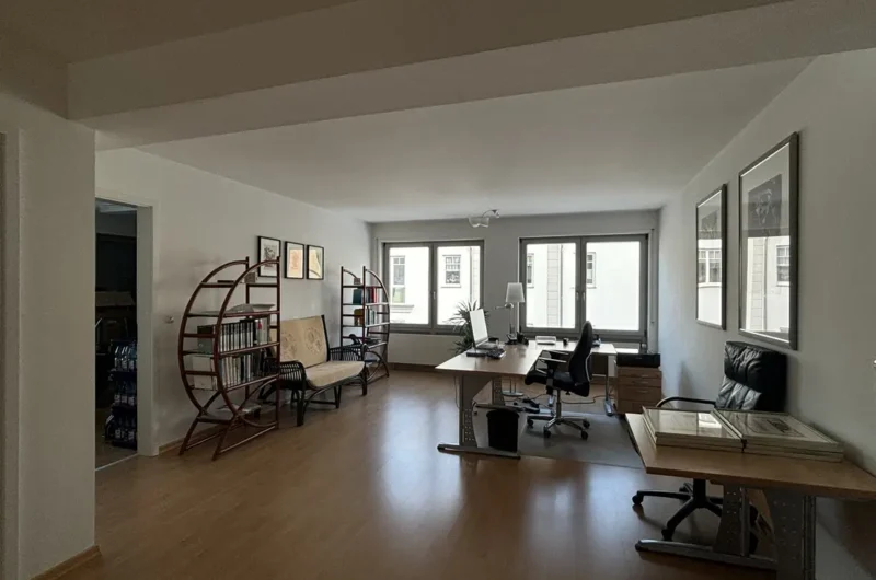 Spacious home office with a desk and computer near large windows, a black swivel chair, filing drawers, and framed art on white walls.