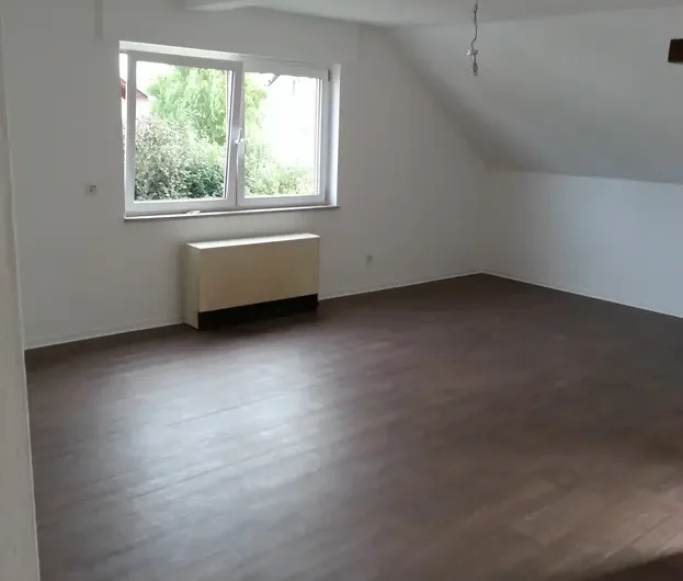 Bright empty attic room with a sloped ceiling, large window, radiator under the window, and dark wood flooring.