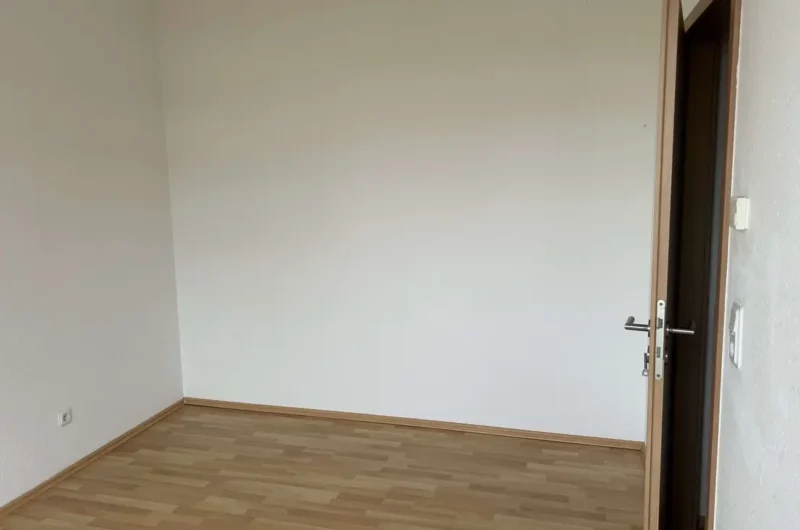 Empty room with light wood flooring and plain white walls; an open wooden door on the right. A power outlet is visible near the left wall.