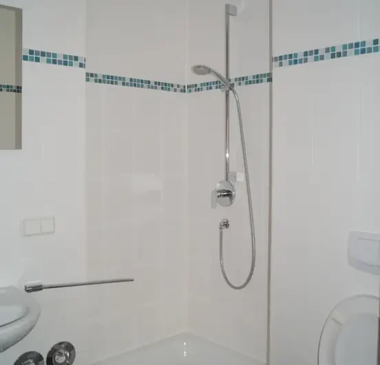 White bathroom featuring a corner shower with handheld shower, blue mosaic border, a nearby toilet and sink.