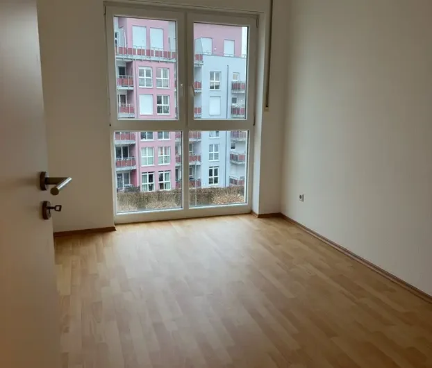 Empty, sunlit room with light wooden floor and a large window overlooking neighboring pink and blue apartment buildings; door partly open on the left.