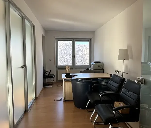 Narrow office with frosted glass doors on the left, a central desk facing a window, black leather chairs, and a printer against the far wall.