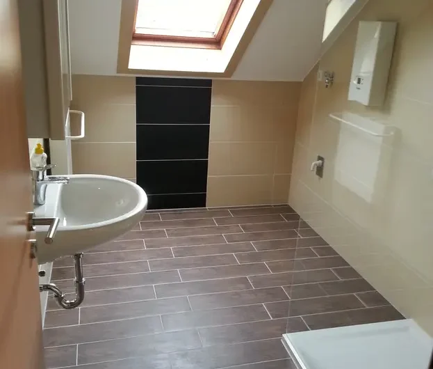 Small bathroom with a skylight, beige wall tiles, and a white sink; dark brown rectangular floor tiles and a corner shower tray visible in the room.