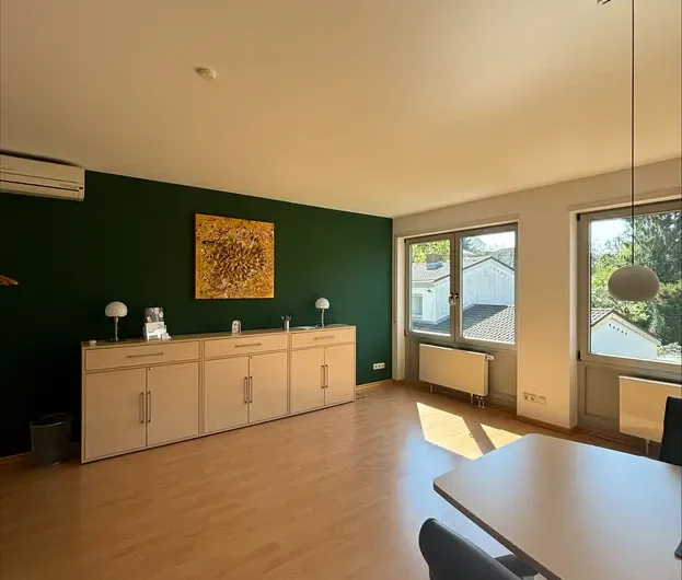 Bright open living area with a dark green accent wall, white sideboard, and sunlight streaming through large windows.