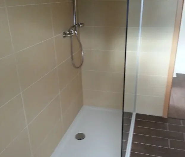 Beige-tiled shower with glass door, chrome handheld shower, and white rectangular base with a round drain effect in a bathroom corner.