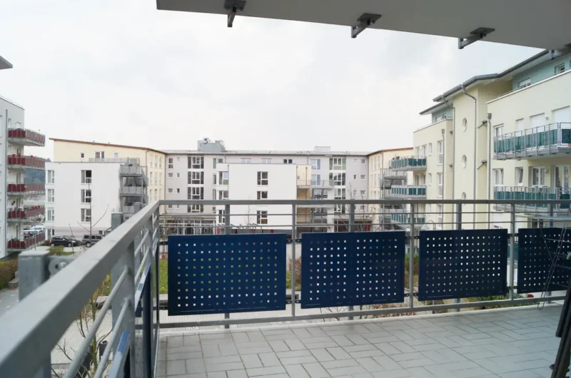 Balcony railing with blue perforated panels overlooking a modern apartment complex courtyard under an overcast sky